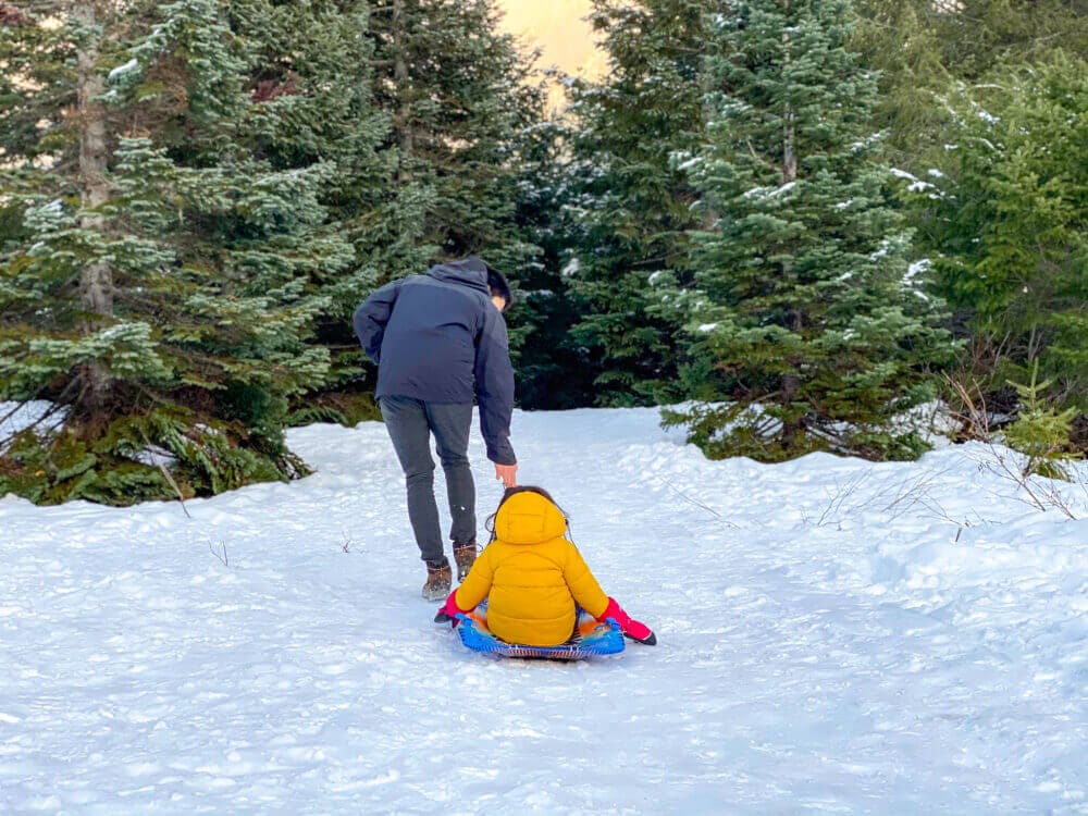 Dad pulling his bundled-up child on a sled along a snow-covered trail at Gold Creek Pond during winter in Snoqualmie Pass. Surrounded by snowy trees and stunning mountain views, this scenic and family-friendly destination offers flat, easy trails perfect for winter walks, snowshoeing, and sledding with young kids—making it a peaceful escape for families near Seattle.