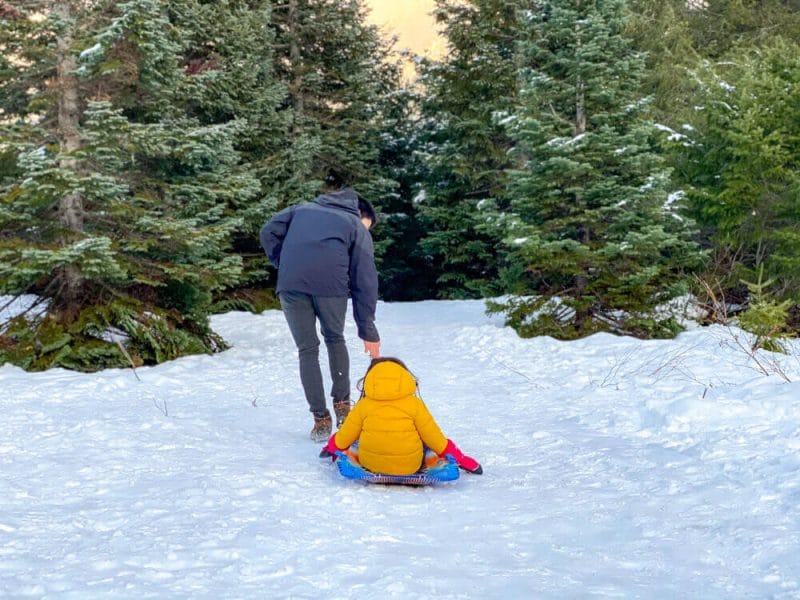 Dad pulling his bundled-up child on a sled along a snow-covered trail at Gold Creek Pond during winter in Snoqualmie Pass. Surrounded by snowy trees and stunning mountain views, this scenic and family-friendly destination offers flat, easy trails perfect for winter walks, snowshoeing, and sledding with young kids—making it a peaceful escape for families near Seattle.