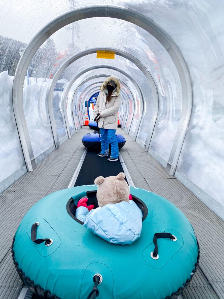 Mom and child riding together in a snow tube on the covered magic carpet lift at The Summit at Snoqualmie tubing hill. This easy, hands-free lift carries families back up the hill after each exhilarating ride, making it fun and accessible for kids and adults. The tubing area is one of the most popular winter attractions near Seattle, offering snowy excitement with minimal effort.