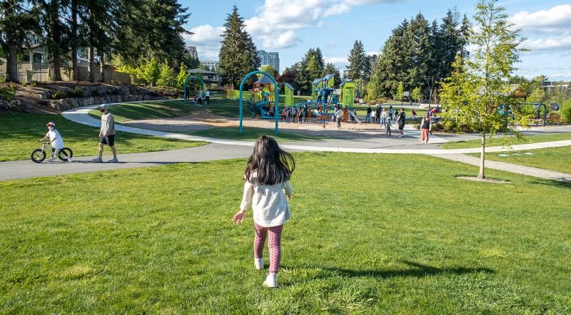 Toddler girl running across the open grassy fields toward the playground at Surrey Downs Park in Bellevue, Washington. This family-friendly park features wide open spaces, shaded picnic areas, and a well-designed playground with structures for both toddlers and older kids—making it a great destination for outdoor play and gatherings on the Eastside.