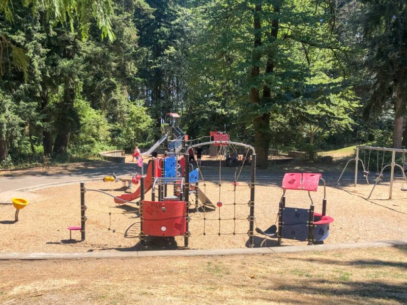 Children enjoying the playground at Northacres Park in North Seattle on a sunny day, with colorful play structures, swings, and tall trees surrounding the area.