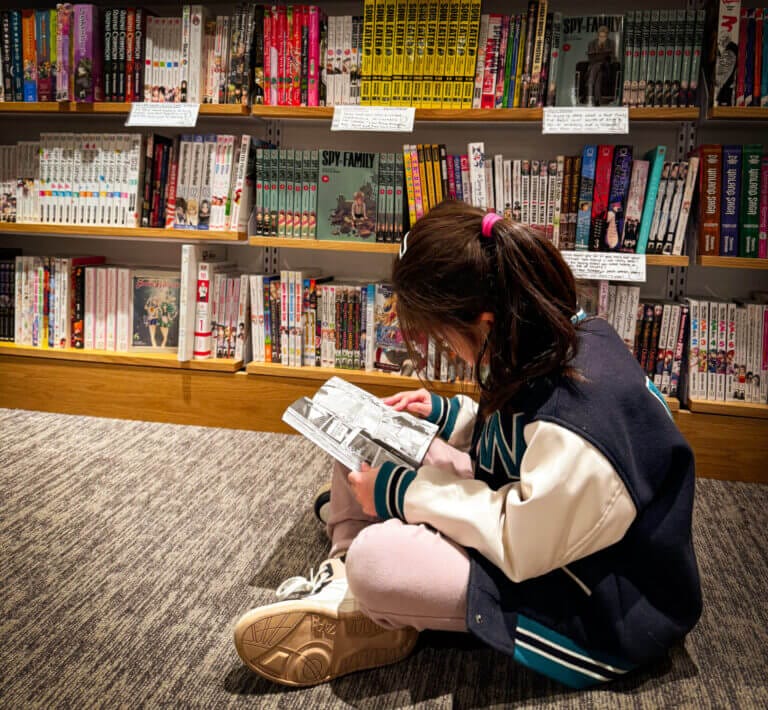 Elementary school girl sitting on the ground reading graphic novel manga in front of a book shelf full of popular manga books for kids like Spy X Family