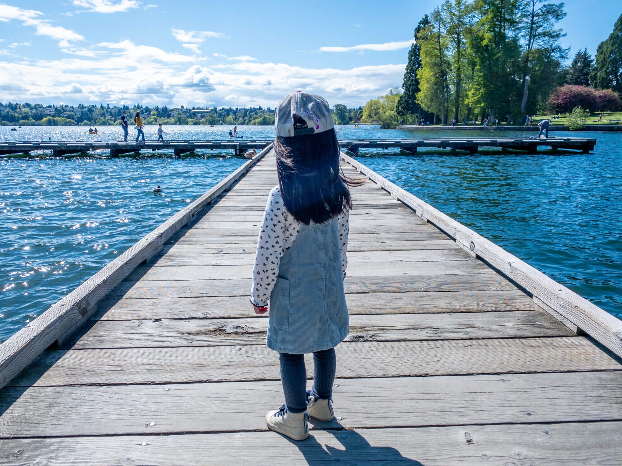 Toddler girl on a dock at Green Lake Park in Seattle, looking out at the water views on a sunny day, with kids paddle boarding in the distance.