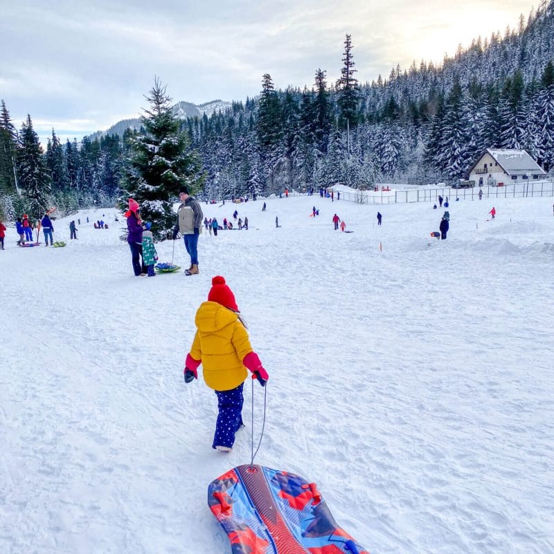Toddler girl bundled in winter gear pulling her sled across the snow at Hyak Sno-Park in Snoqualmie Pass, heading toward one of the many gentle sledding hills. This family-friendly snow park is a favorite for young children and parents, offering designated sledding areas, groomed trails, restrooms, and easy access for a fun winter day in the mountains.
