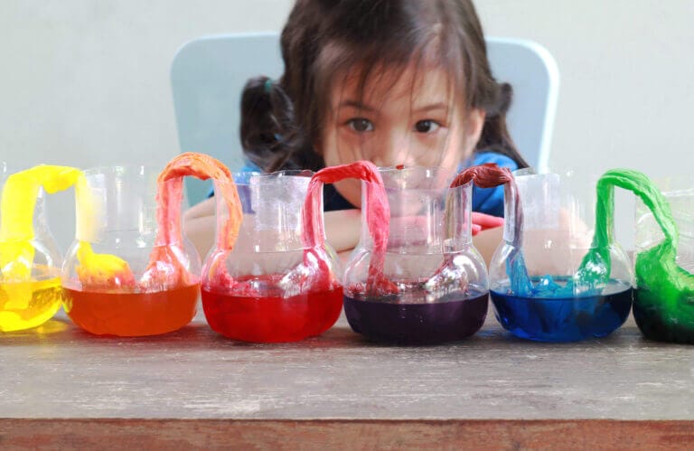 Young girl doing a STEM experiment, looking at glasses with different colored liquids arranged in rainbow colors.