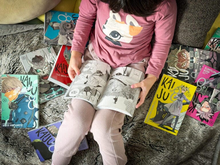 Elementary school girl reading Kaiju No. 8 manga on the floor, surrounded by manga books.