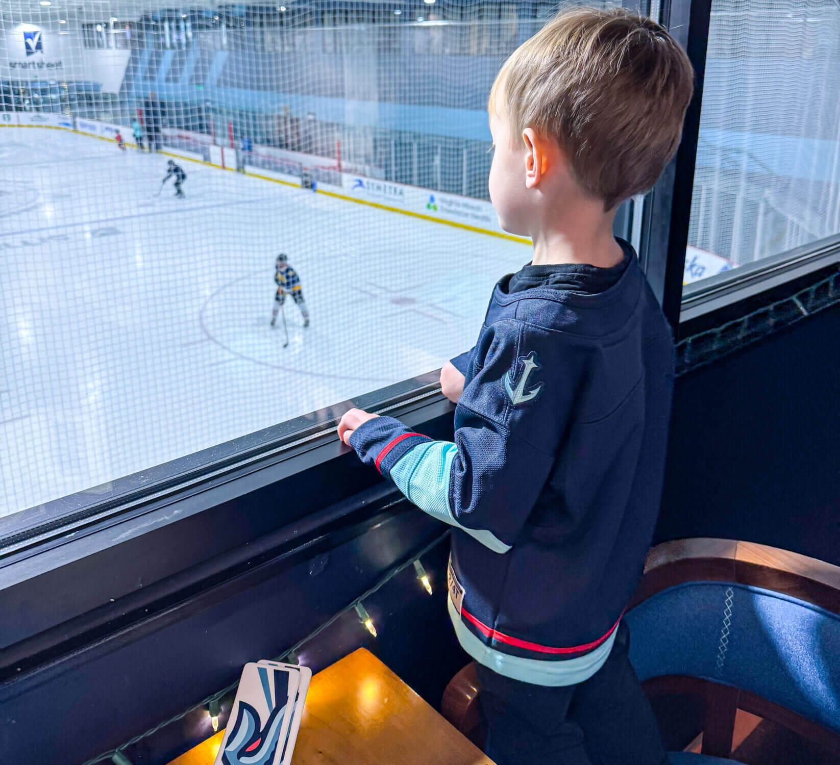 A young boy watches the Seattle Kraken’s team practice session through the large windows of 32 Bar & Grill at the Kraken Community Iceplex, all while enjoying a meal. This family-friendly restaurant offers a unique dining experience with rink-side views, allowing fans to watch their favorite NHL team train while savoring delicious food. A perfect spot for hockey enthusiasts and families looking to combine great eats with an up-close look at the Kraken in action.