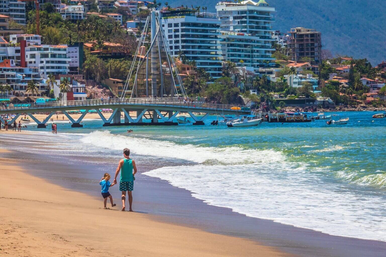 A father and his young daughter walk hand-in-hand along the sandy beach in Puerto Vallarta, Jalisco, with the famous Malecon boardwalk and colorful buildings in the background. This vibrant, family-friendly destination offers beautiful ocean views, lively street art, sculptures, and a relaxed atmosphere perfect for exploring with kids. The scene captures the heart of a beachside stroll in Puerto Vallarta—where family moments and the charm of the city meet