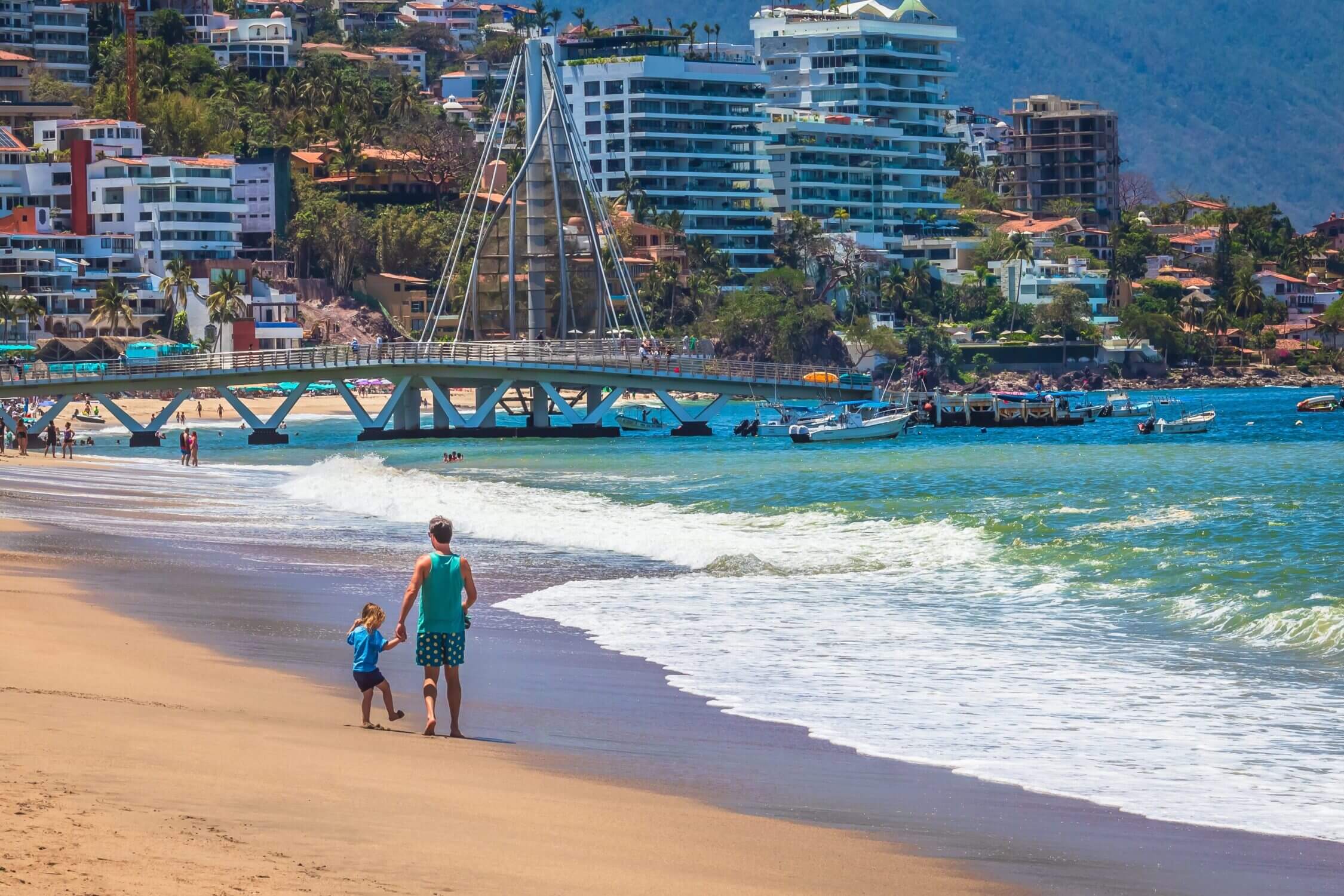 A father and his young daughter walk hand-in-hand along the sandy beach in Puerto Vallarta, Jalisco, with the famous Malecon boardwalk and colorful buildings in the background. This vibrant, family-friendly destination offers beautiful ocean views, lively street art, sculptures, and a relaxed atmosphere perfect for exploring with kids. The scene captures the heart of a beachside stroll in Puerto Vallarta—where family moments and the charm of the city meet