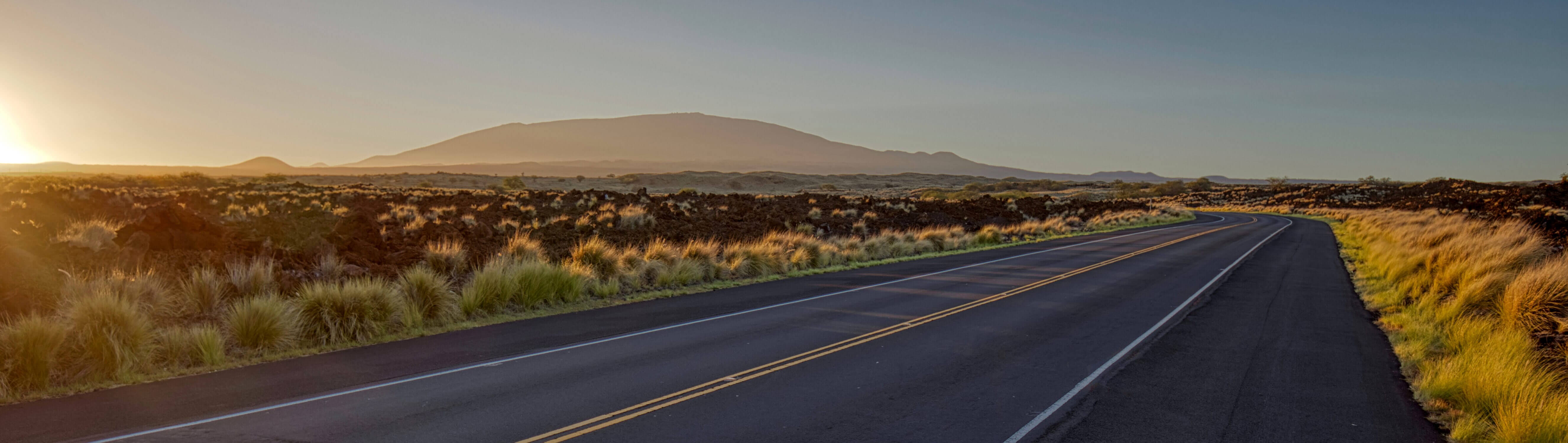 Early morning drive on the Big Island of Hawaii, with a long stretch of empty road surrounded by dramatic black lava rock fields under a glowing sunrise sky. A distant mountain rises on the horizon, capturing the raw beauty of the island’s landscape. Featured in our blog about doing a day trip from Kona to Hilo, this scenic route is a highlight in itself—offering peaceful, otherworldly views and a unique driving experience between two sides of the island.