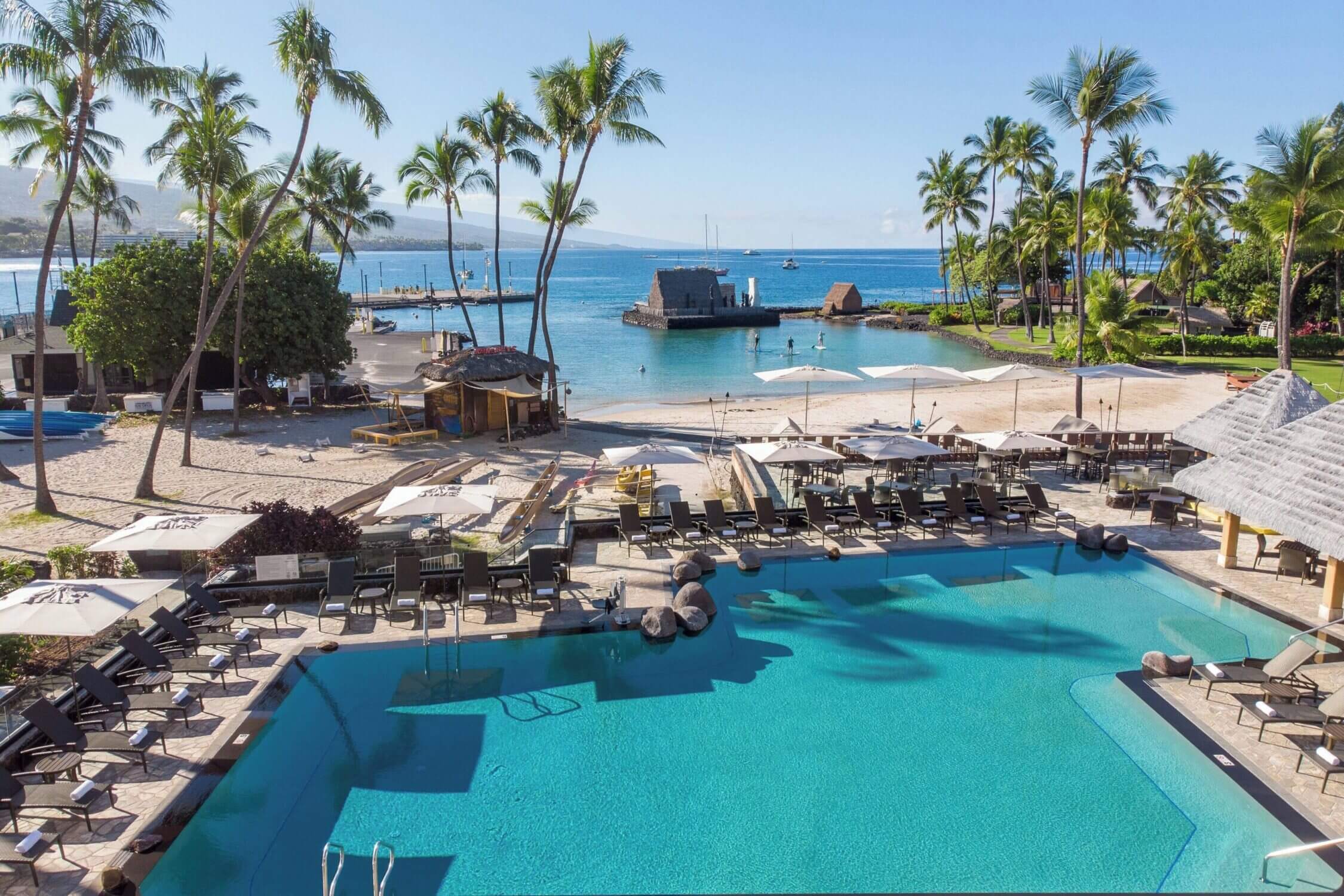 Aerial view of the pool area at the Courtyard by Marriott King Kamehameha’s Kona Beach Hotel on the Big Island, with a stunning backdrop of the calm, sandy beach and clear ocean waters. This centrally located family-friendly resort in Kailua-Kona offers easy beach access, kid-friendly swimming areas, and walkable proximity to local shops, historic sites, and restaurants—making it a top pick for families visiting Kona.
