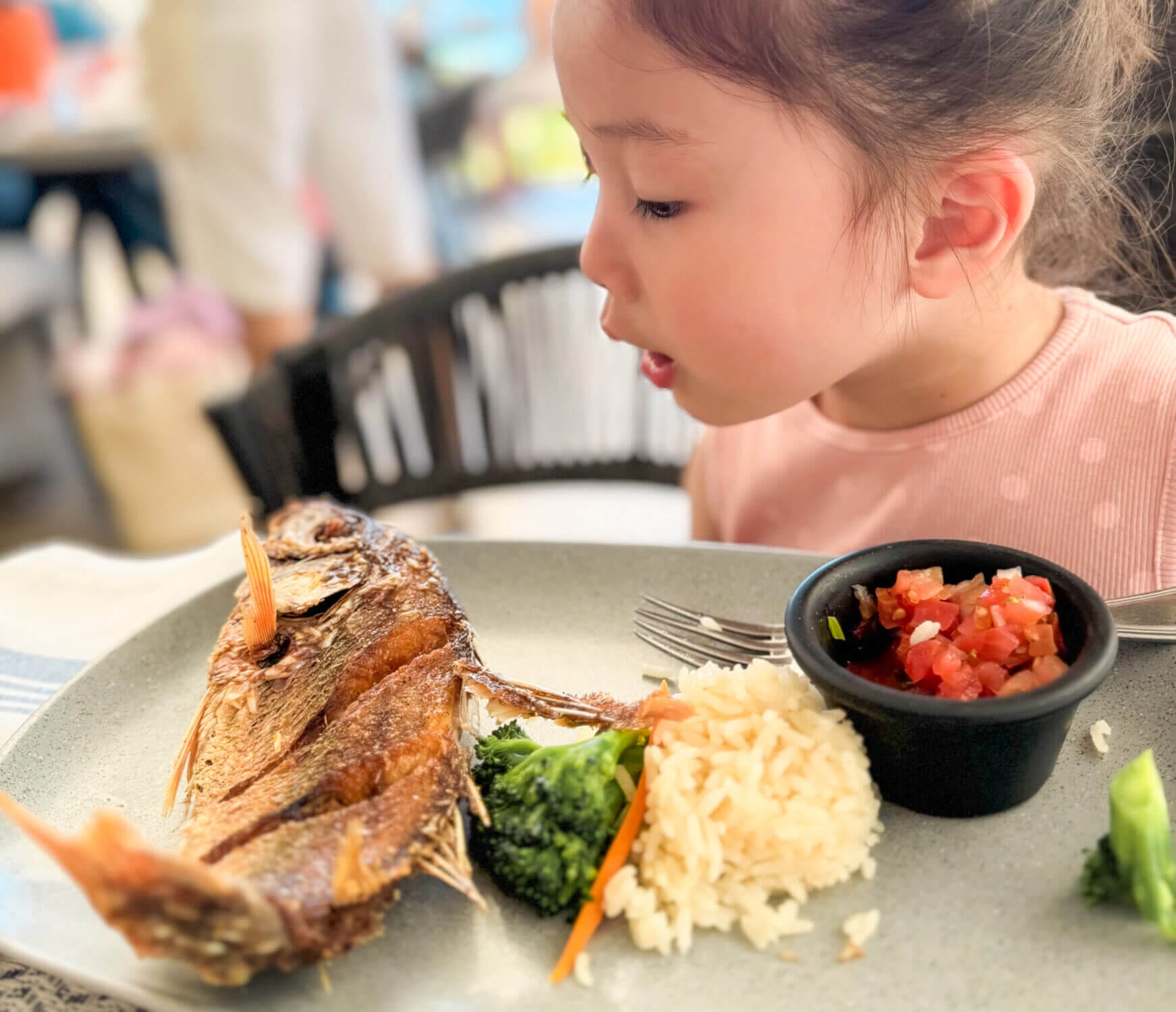 A toddler girl curiously examines her fried whole fish dinner at Castaways, the open-air beachfront restaurant at Dreams Vallarta Bay Resort. This all-inclusive dining experience offers fresh seafood and ocean views, with kid-friendly options and adventurous dishes alike. It’s a great place for families to enjoy a relaxed dinner after a day of fun at the pool or beach