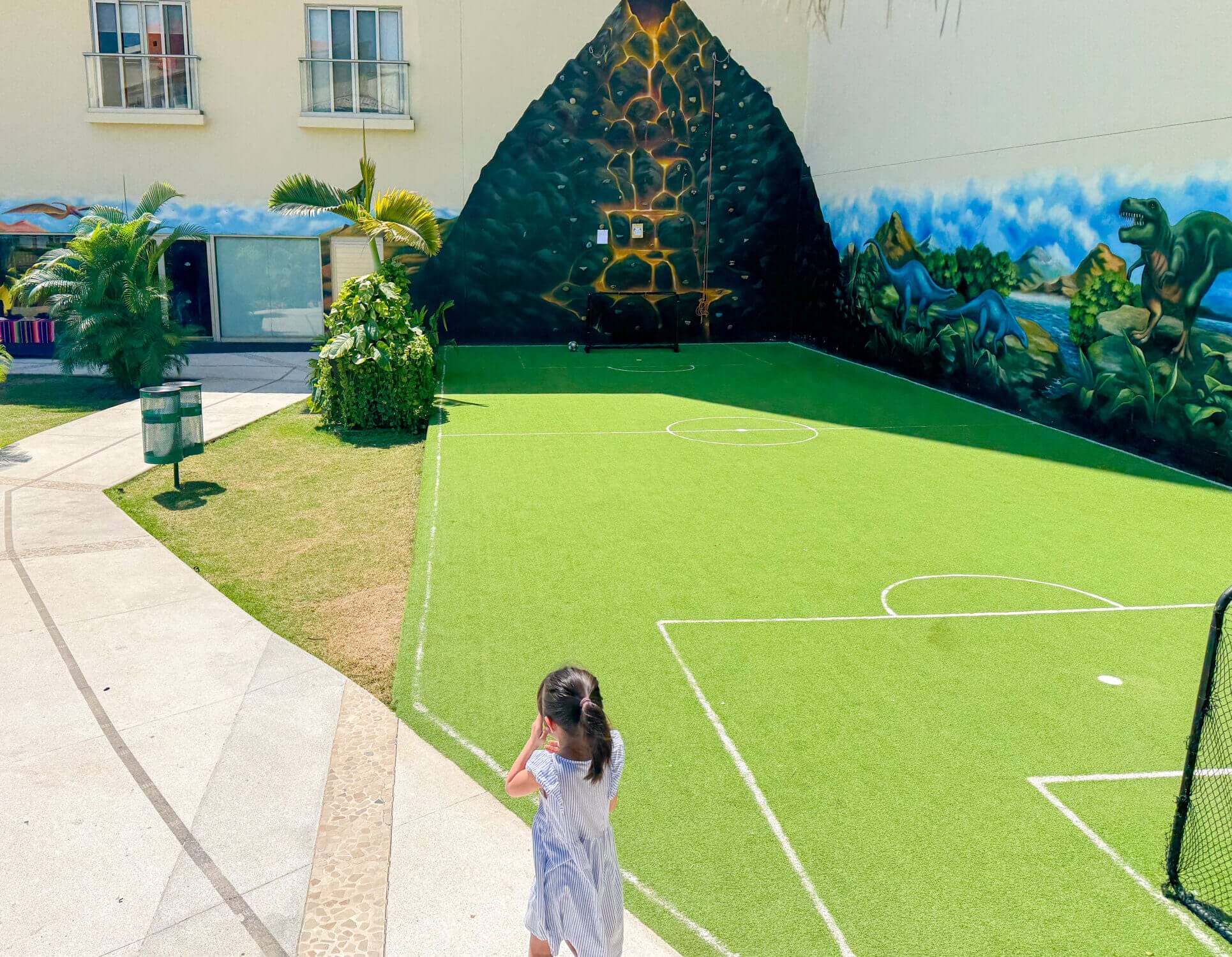 A young girl walks toward the indoor section of the Dreams Vallarta Bay Resort Kids Club, where children can color, play games, and enjoy air-conditioned fun. On her way, she passes the outdoor play area featuring a mini soccer field and kid-sized rock climbing wall—both popular activities among young guests. This supervised club offers a mix of creative play and outdoor adventure, giving kids a space to make friends while parents enjoy a little downtime at this family-friendly all-inclusive resort