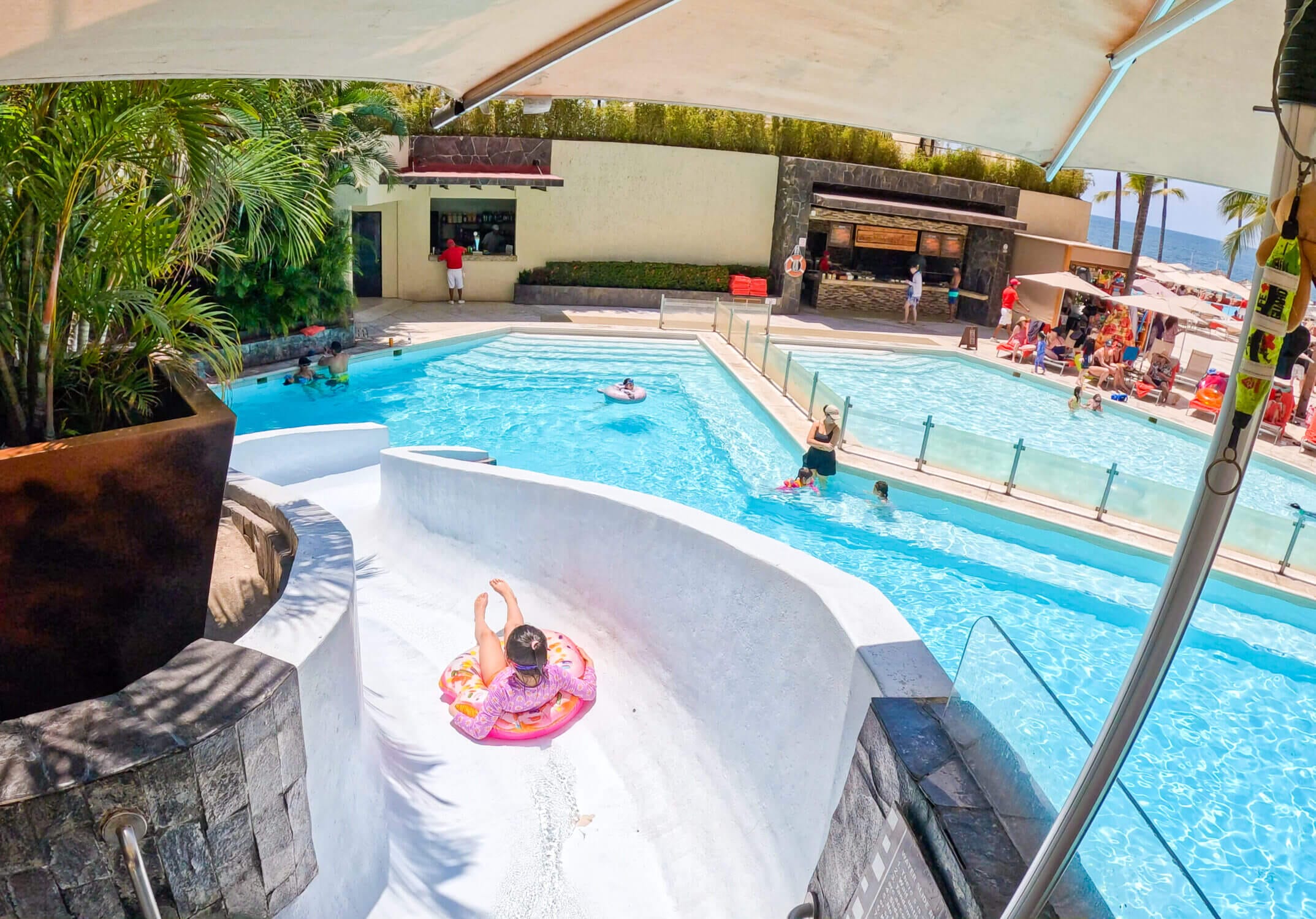 A young girl rides down the water slide on an inflatable at Dreams Vallarta Bay Resort, surrounded by palm trees and vibrant blue pool water. This family-friendly resort in Puerto Vallarta offers the perfect mix of fun and relaxation, with kid-approved water play areas, stunning ocean views, and all-inclusive amenities. The poolside excitement makes it an ideal destination for family vacations, where kids can splash all day while parents unwind in tropical paradise.