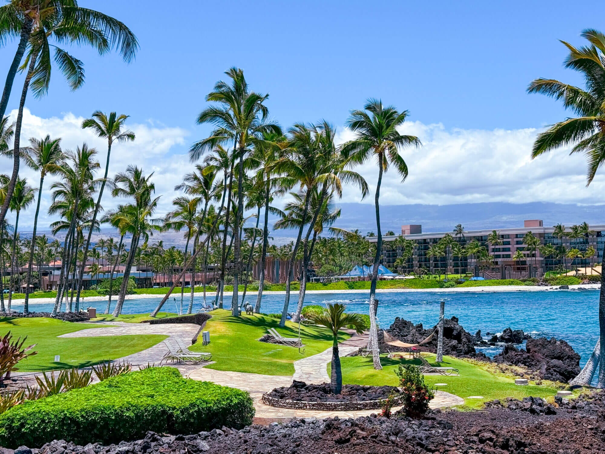 Scenic view from Hilton Waikoloa Village near Buddha Point on the Big Island, with dramatic black lava rocks in the foreground, the sparkling blue ocean beyond, and parts of the resort visible in the background. The resort’s lush walking paths wind through art, sculptures, and natural landscapes, offering a peaceful and family-friendly way to explore Kona’s unique beauty—even if you’re just visiting for the day.