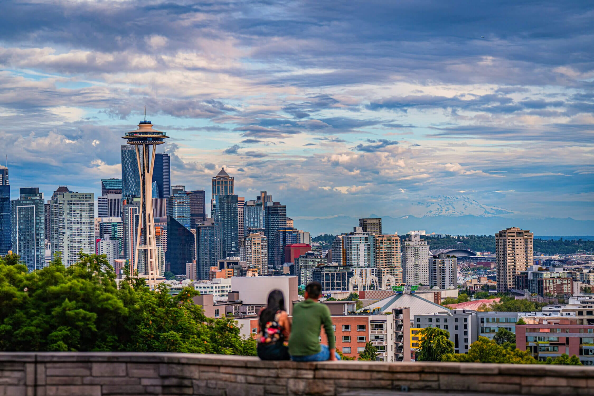Sunset view from Kerry Park in Seattle with a couple sitting on the lookout wall, softly blurred in the foreground, as the Space Needle and city skyline glow in the background. This iconic viewpoint offers breathtaking vistas and is a must-see destination for anyone visiting Seattle, especially during golden hour.