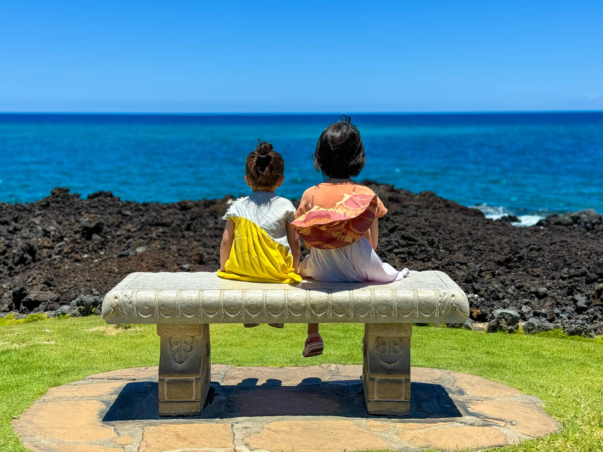 Two little sisters sit on a bench at the Hilton Waikoloa Resort on the Kona side of the Big Island of Hawaii, gazing out at the brilliant blue ocean with rugged black lava rocks in the foreground. This serene family moment highlights the beauty of Kona’s sunny coastline and is part of a blog comparing Hilo vs. Kona to help families decide which side of the island is best for their vacation.