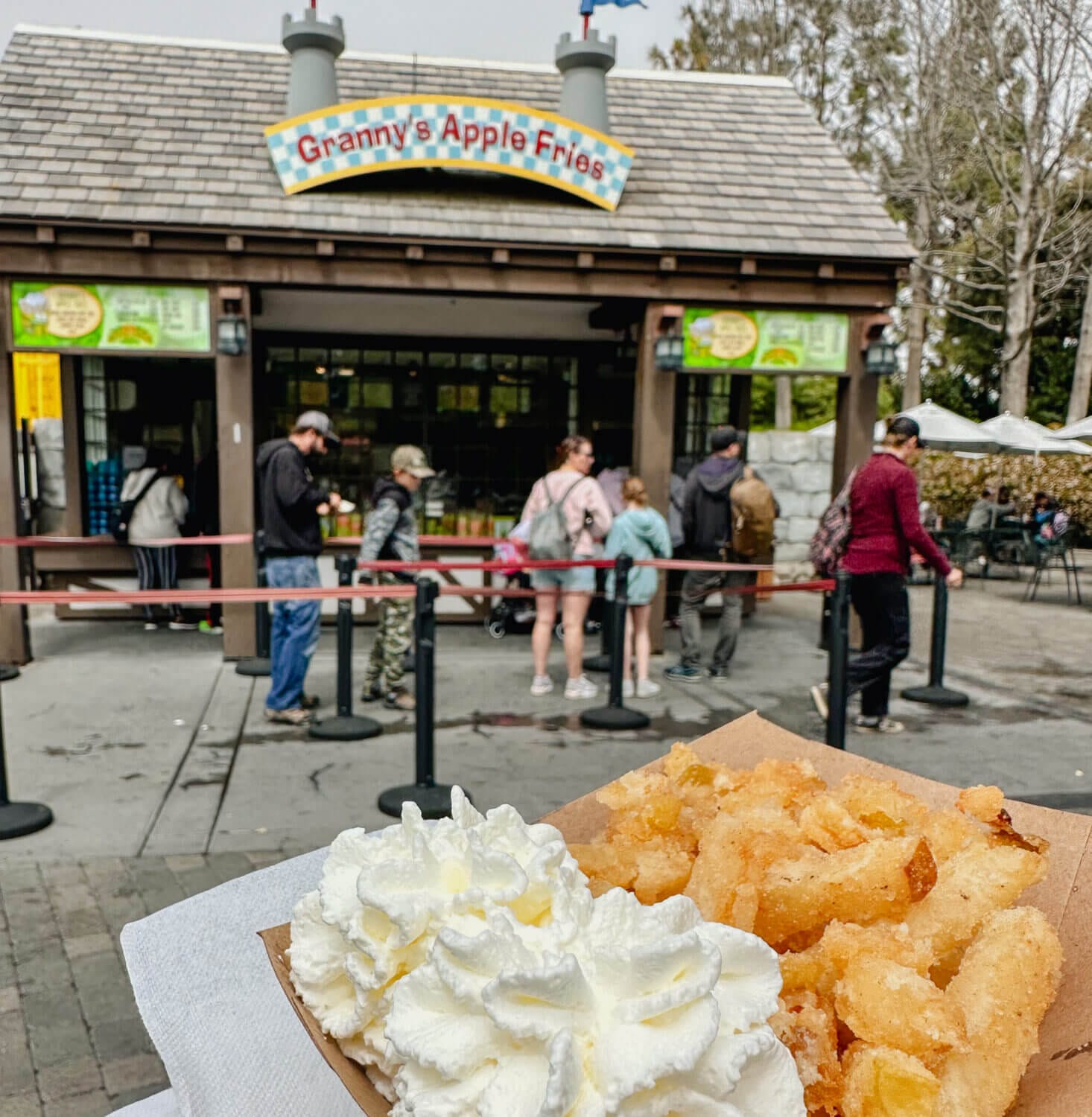 Close-up of a hand holding a serving of Granny’s Apple Fries at LEGOLAND California, with the background blurred to show the signature stand behind. These warm, cinnamon-sugar coated apple fries served with a sweet whipped cream dip are one of the park’s most iconic and beloved snacks, making them a must-try treat featured in our LEGOLAND California family guide.
