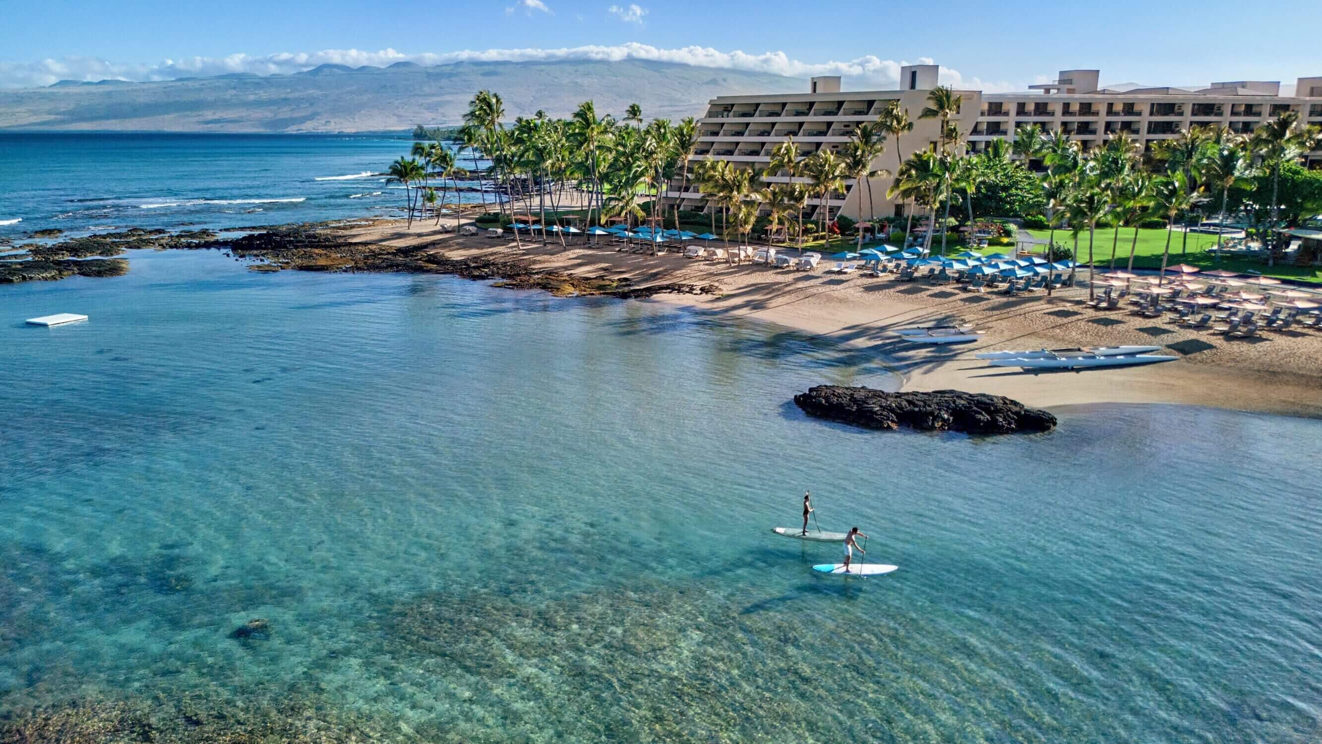 Aerial view of the Mauna Lani resort on the Kona side of the Big Island, featuring the elegant hotel grounds, white sand beach, and calm turquoise waters where two people are paddleboarding. Highlighted as one of our top luxury picks for families, Mauna Lani offers a serene and upscale experience with family-friendly amenities, cultural activities, and easy beach access—perfect for a relaxing and memorable Hawaii vacation.