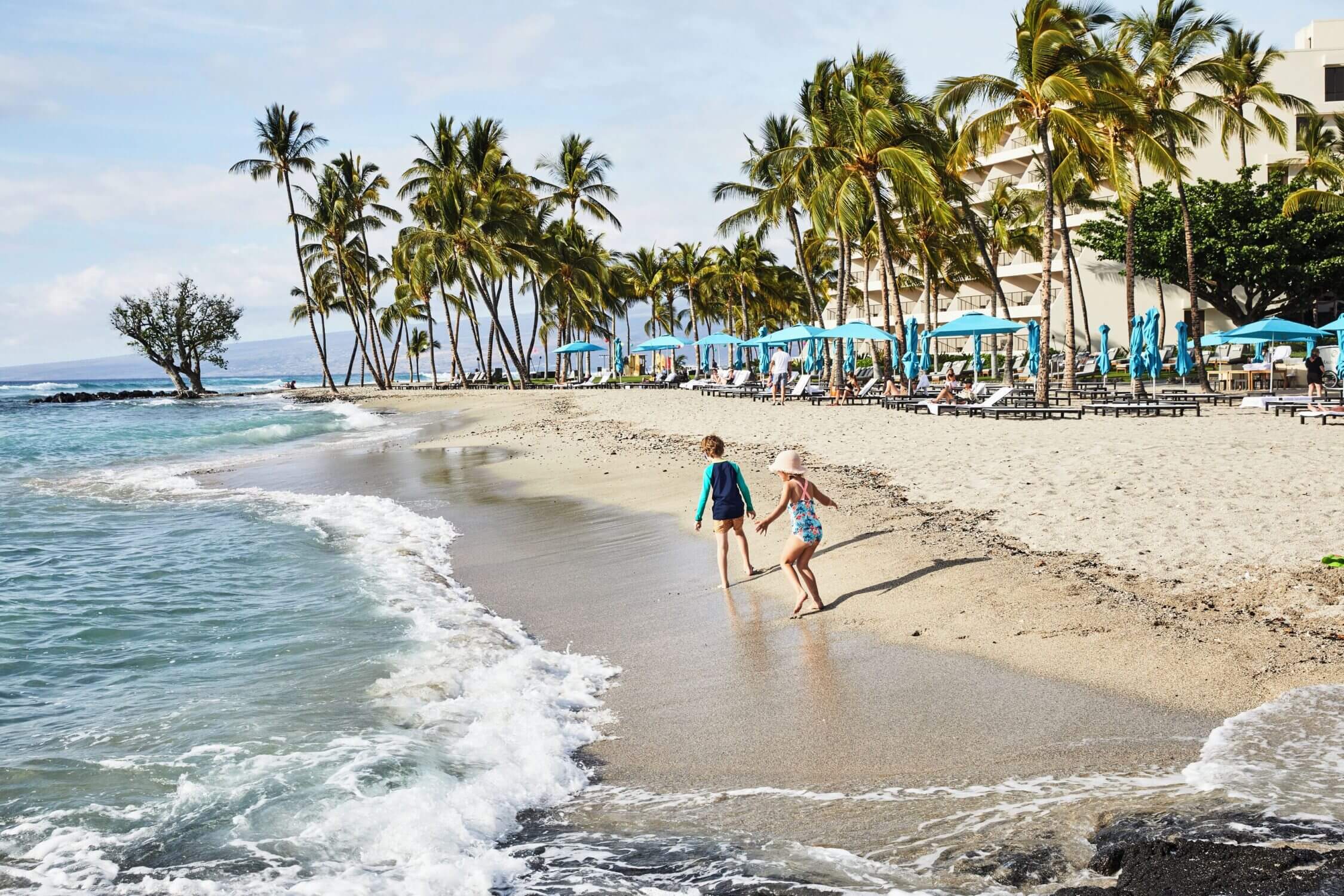 Two young kids walking along the shoreline at Mauna Lani Beach on the Kona side of the Big Island, with soft white sand beneath their feet. The beach is lined with shaded cabanas and sun loungers, offering a relaxing and upscale setting for families. As one of our luxury resort picks, Mauna Lani provides calm waters, cultural experiences, and a peaceful beachfront—ideal for family getaways in Hawaii