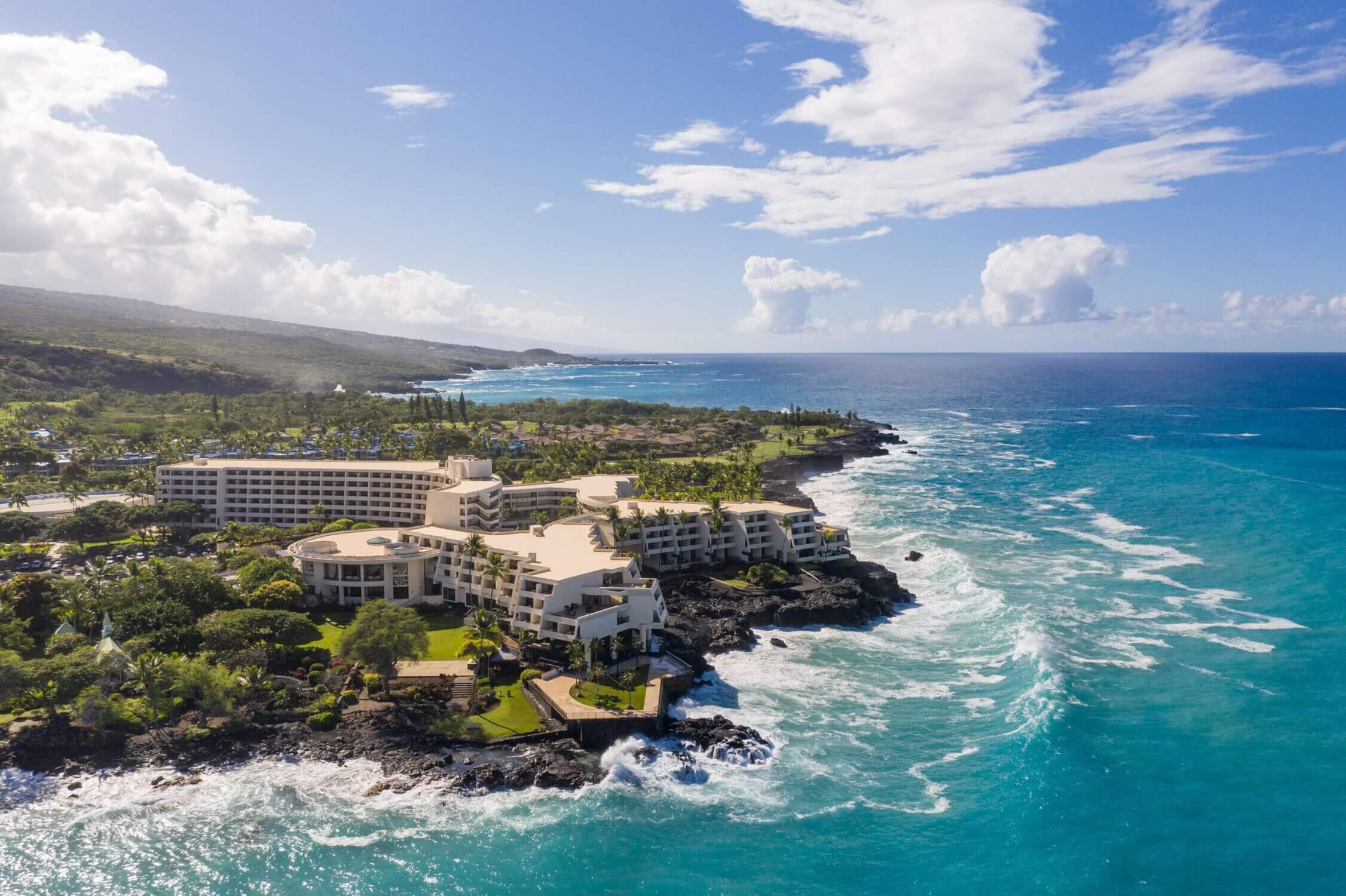 Aerial view of the Outrigger Kona Resort on the Big Island of Hawaii, perched dramatically on a rocky ocean shoreline with stunning coastal views. Featured in our roundup of best resorts for families in Kona, this resort offers spacious rooms, a kid-friendly pool with a waterslide, cultural experiences, and on-site luau—making it a top pick for an unforgettable family vacation.