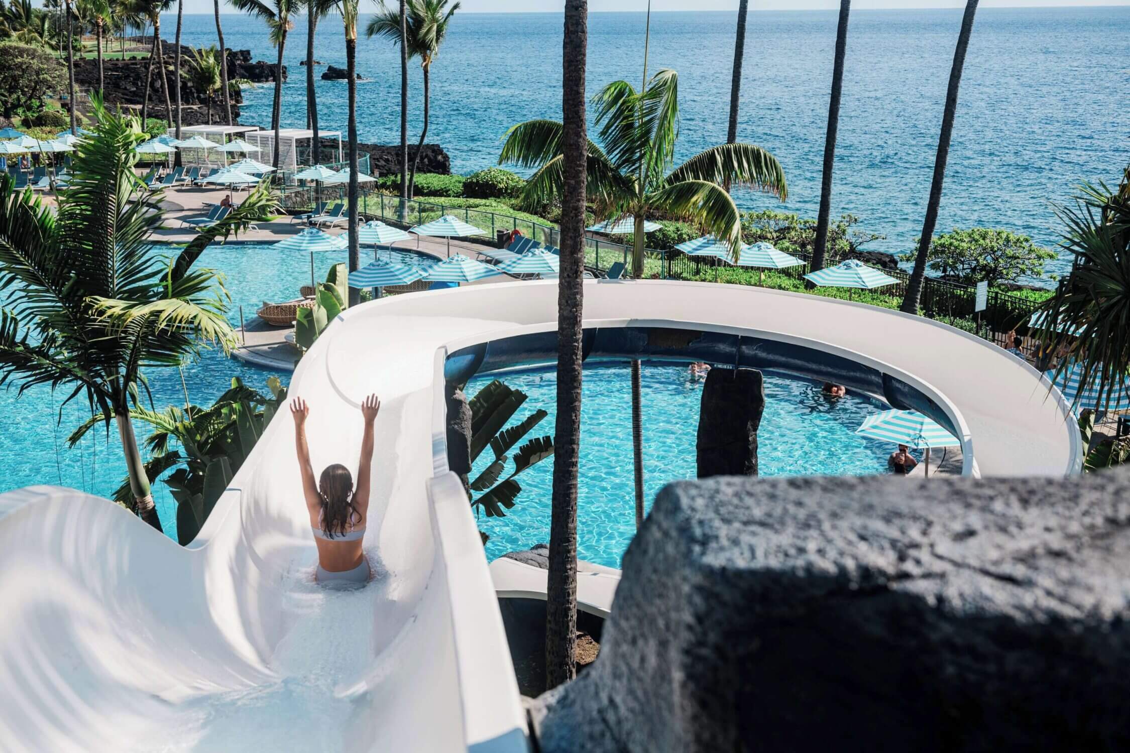 Child sliding down the waterslide at the Outrigger Kona Resort on the Big Island of Hawaii, hands in the air with the sparkling ocean in the background. This kid-friendly pool area is a highlight for families, offering hours of fun in a tropical setting. With stunning oceanfront views and resort amenities designed for all ages, the Outrigger Kona is one of the top family-friendly resorts in Kona.