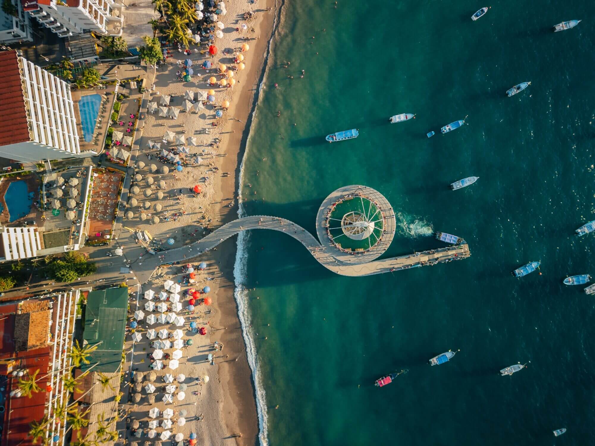 Aerial view above Los Muertos Beach Pier in Puerto Vallarta, capturing the vibrant scene of boats anchored in the bay, beachgoers relaxing under colorful umbrellas, and tourists strolling the iconic pier. This lively and popular tourist spot is a must-visit for families looking for sun, sand, and seaside fun.