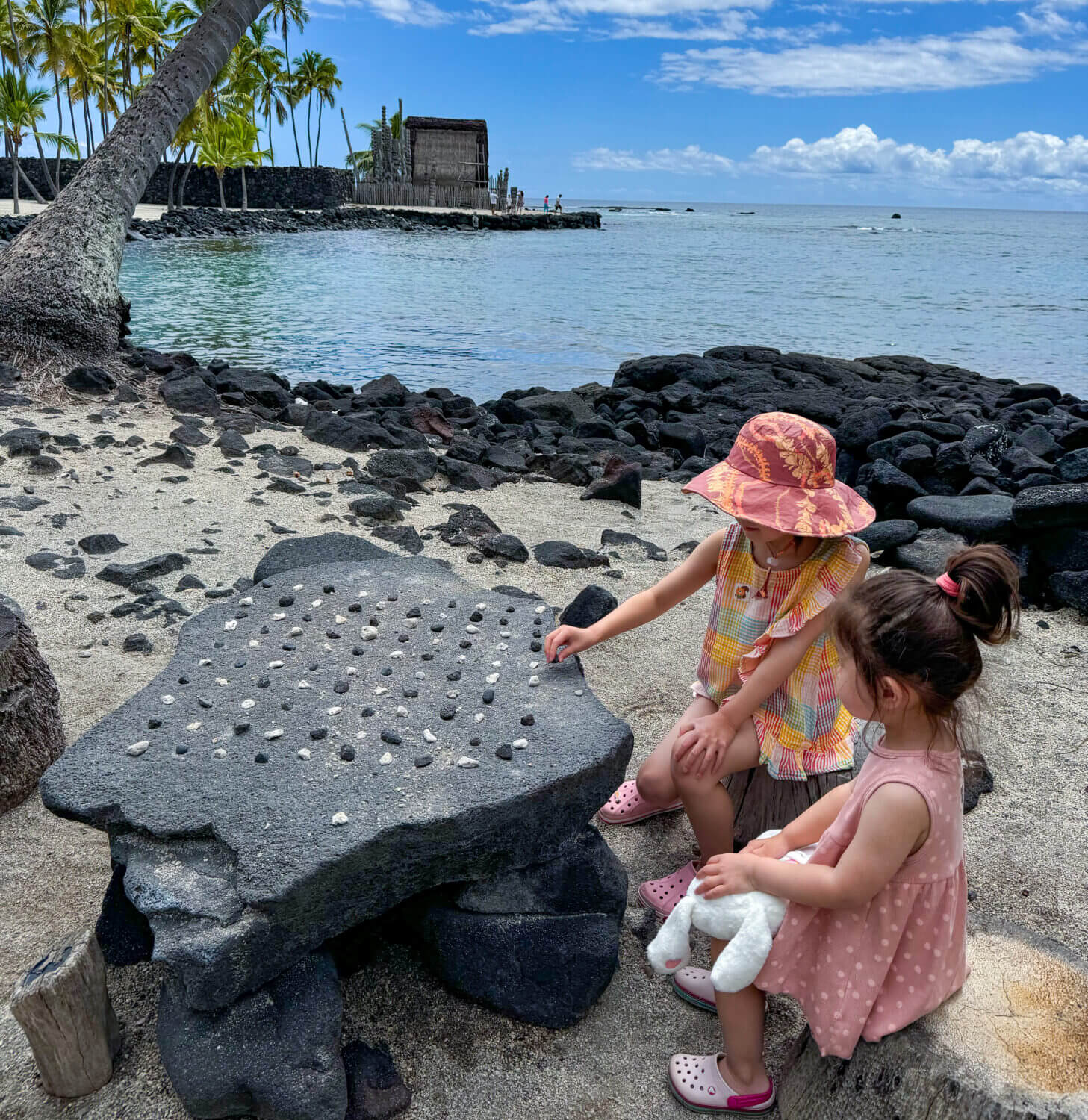Two young kids sitting at a stone game board at Puʻuhonua o Hōnaunau National Historical Park on the Big Island of Hawaii. This rock carving represents a traditional Hawaiian strategy game, offering an interactive and educational moment during a family visit to this sacred site. The park is rich with cultural history, restored temples, and coastal trails—making it both a meaningful and fun place to explore with kids in Kona.