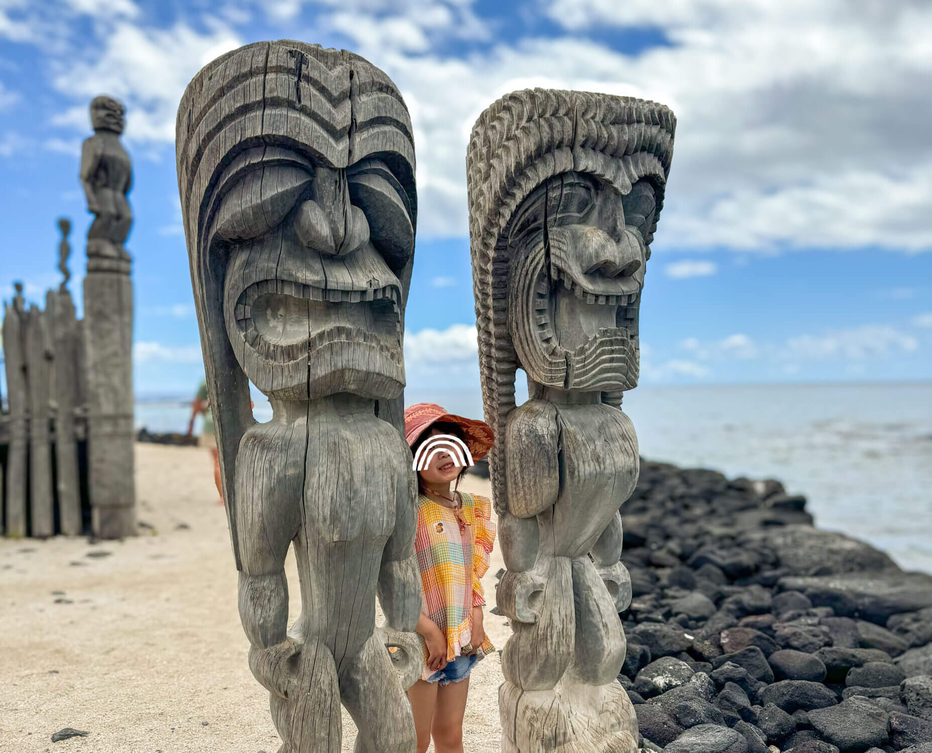 Young girl taking a photo between two traditional tiki statues at Puʻuhonua o Hōnaunau National Historical Park, also known as the City of Refuge, on the Kona side of the Big Island of Hawaii. This sacred site offers a unique cultural and historical experience for families, with preserved temples, wooden carvings, and coastal walking paths—making it a meaningful and educational stop for kids in Kona.