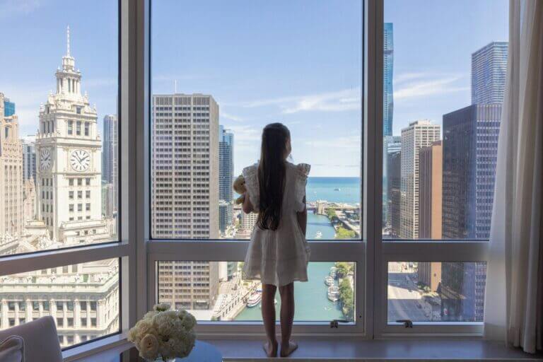 Young girl gazing out the window of a Chicago hotel room, with a scenic view of the Chicago River and iconic downtown architecture—highlighting one of the top kid-friendly hotels featured in our blog.