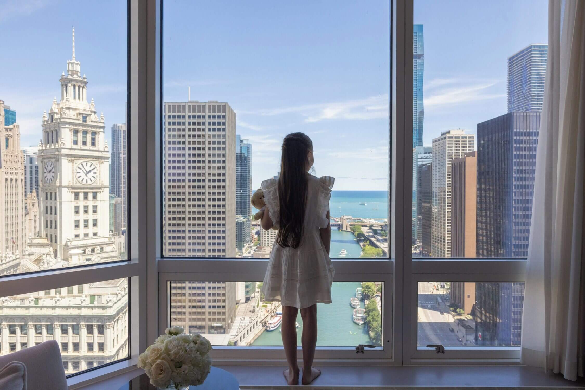 Young girl gazing out the window of a Chicago hotel room, with a scenic view of the Chicago River and iconic downtown architecture—highlighting one of the top kid-friendly hotels featured in our blog.