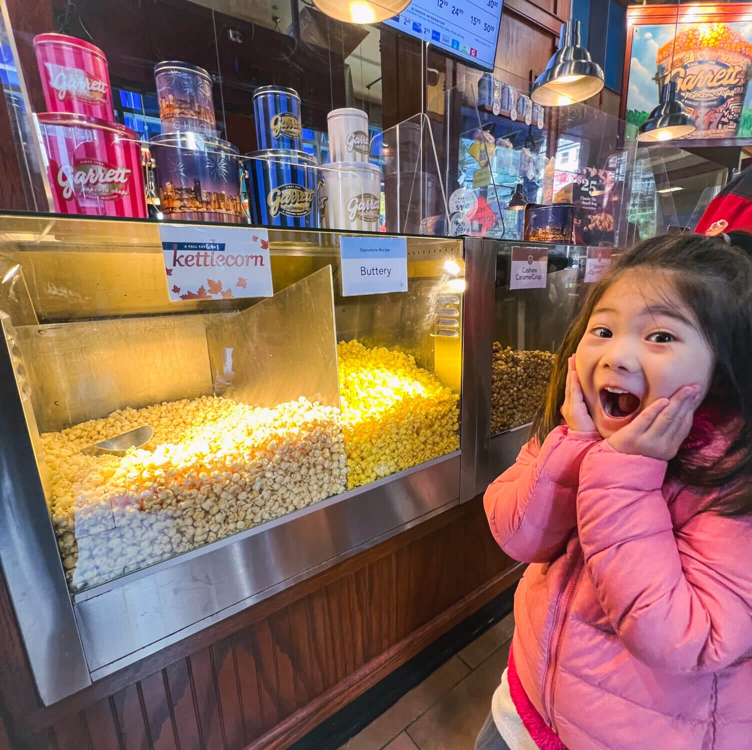 Young girl with an excited expression posing in front of the popcorn display at Garrett Popcorn in Chicago, a must-visit treat for families exploring the city with kids—famous for its Chicago Mix of cheese and caramel popcorn.